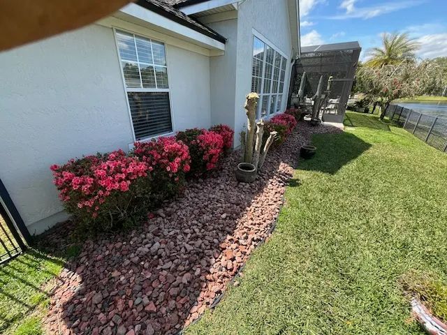 Side yard with white house, pink flowers, red mulch border, green lawn, and patio area beside a canal
