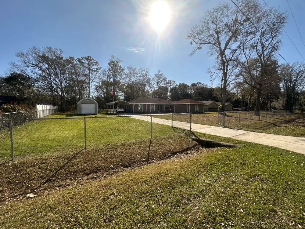 Chain-link fenced yard with a paved path, basketball hoop, and trees in bright sunlight