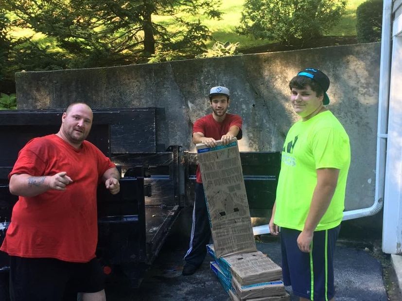 Three people stand by a barbecue grill outdoors; one holds a grill brush and another wears a neon shirt.
