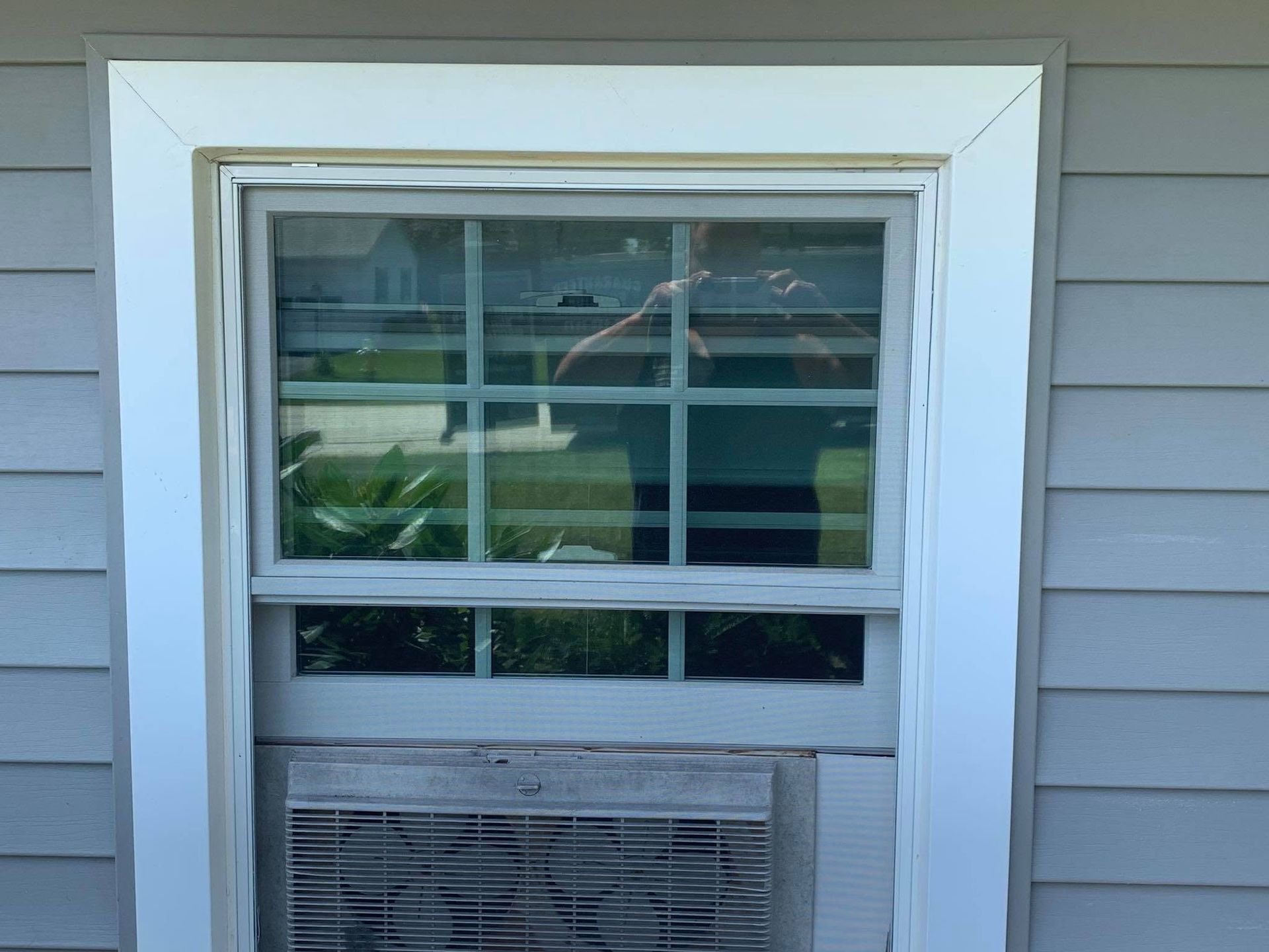 White-framed window in light gray siding with a window AC unit below and blinds inside.