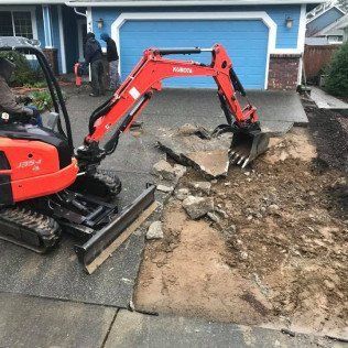 A small excavator is digging a hole in a driveway in front of a house.