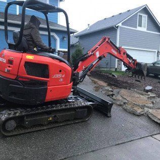 A small red excavator is sitting on the side of the road in front of a house.
