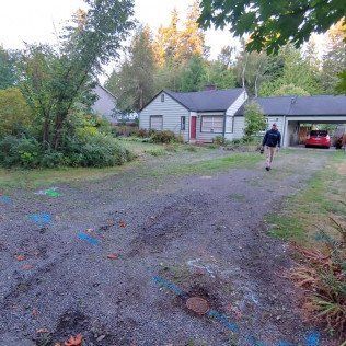 A man is walking down a dirt road in front of a house.