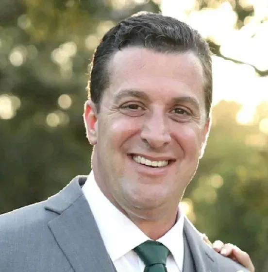 Man in grey suit smiling, with green tie and collar, outdoors with blurred background.