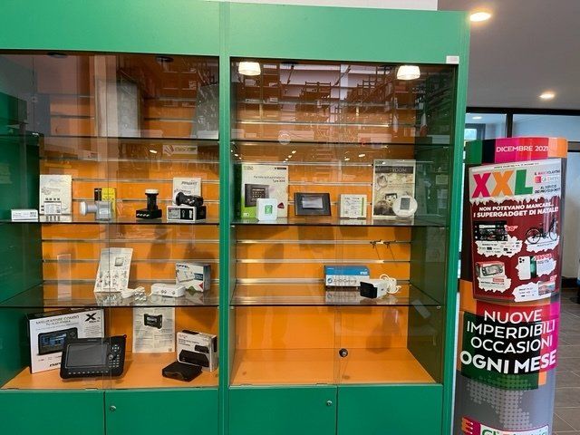 Vetrina che espone prodotti elettronici in un negozio. Un mobiletto verde e arancione contiene vari dispositivi, con una pubblicità circolare sulla destra.
