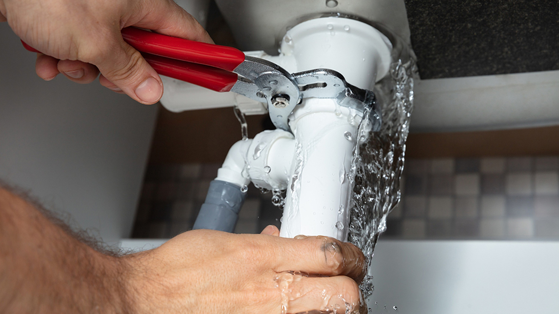 A person uses red-handled pliers to tighten a white plastic sink pipe that is actively leaking water.