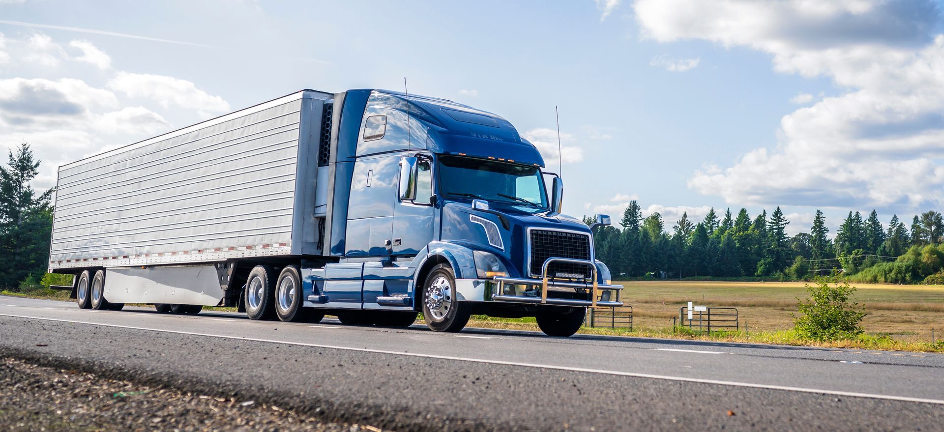 A blue semi truck is driving down a highway next to a field.