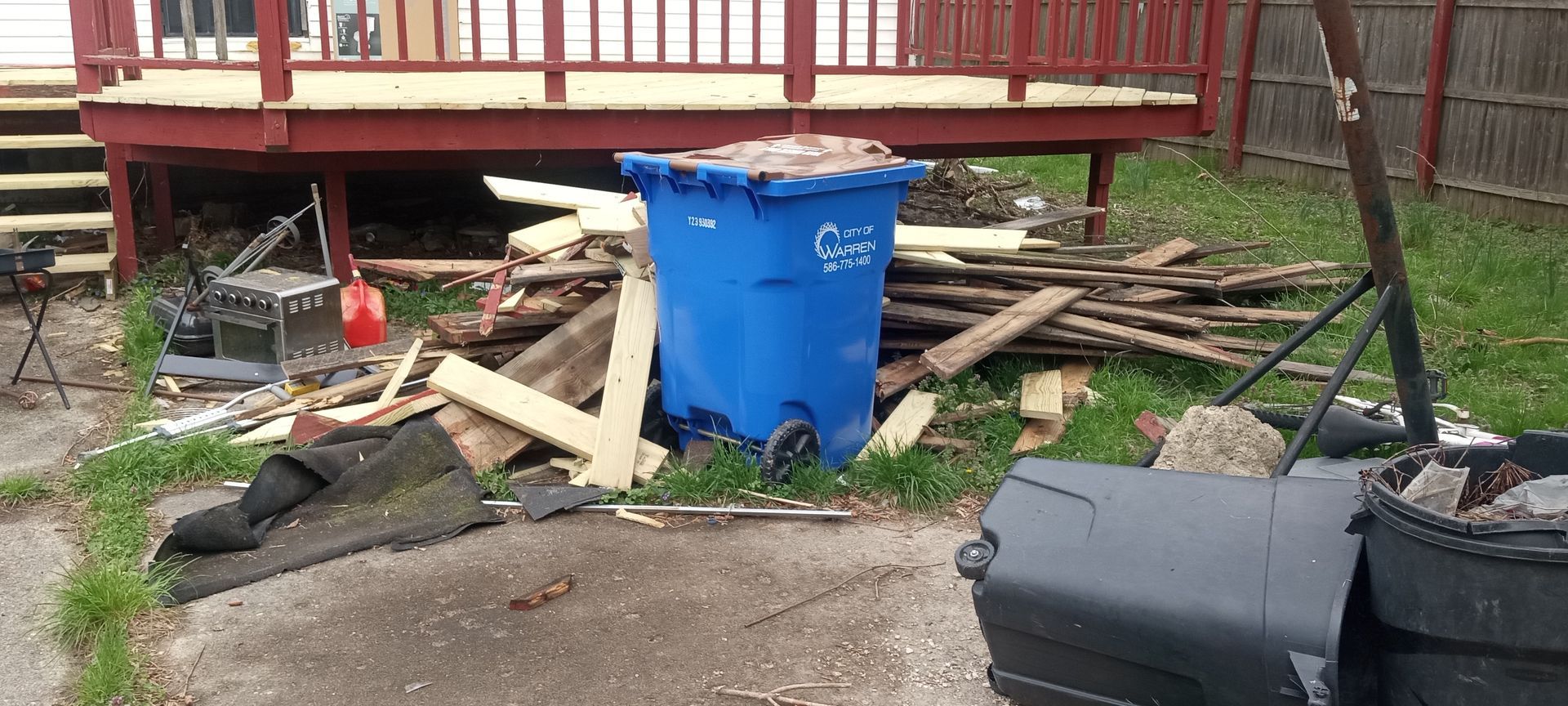 A blue trash can is sitting in front of a pile of wood.