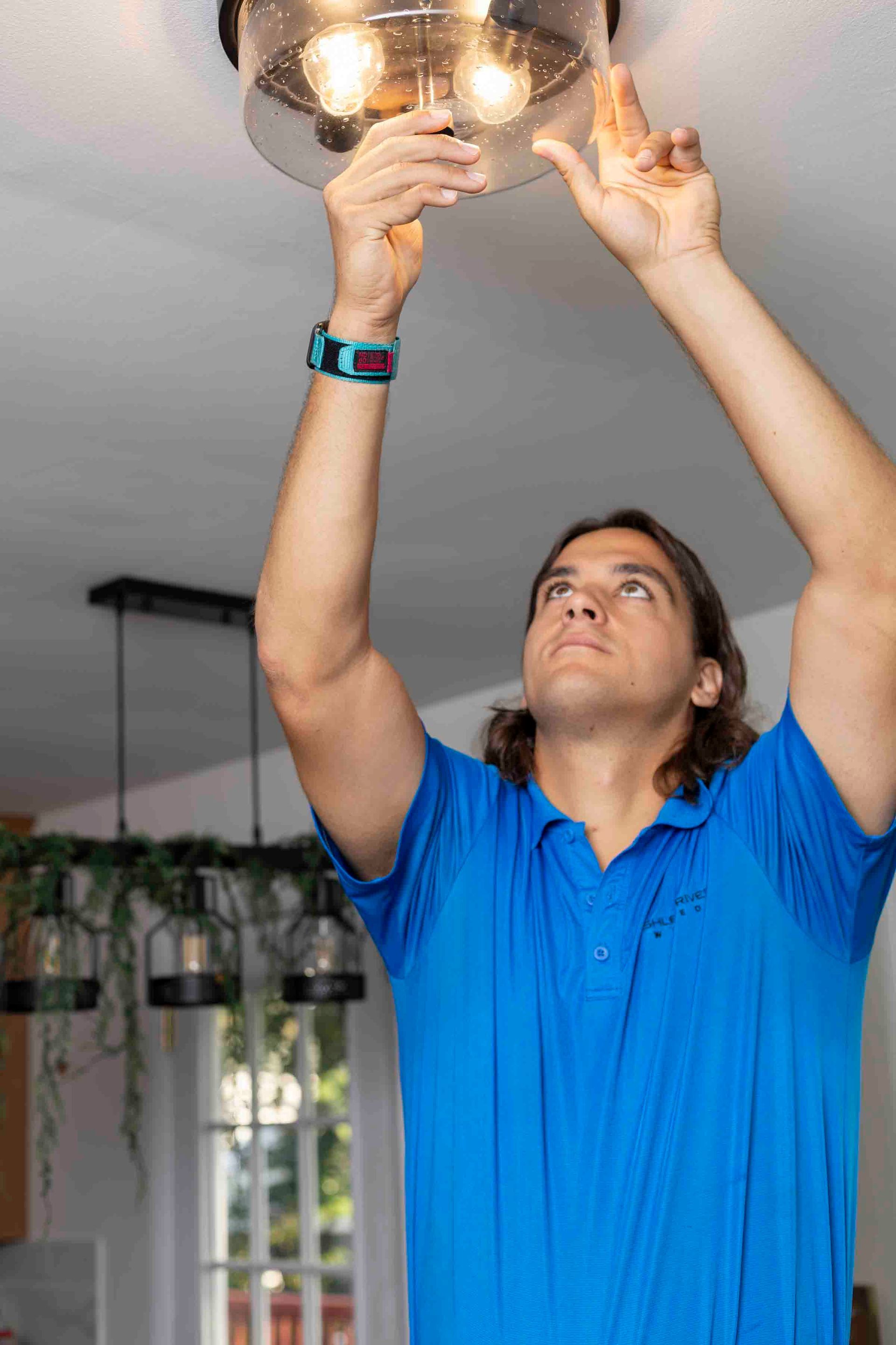 Man in blue shirt changing a ceiling light bulb, reaching up with both hands.