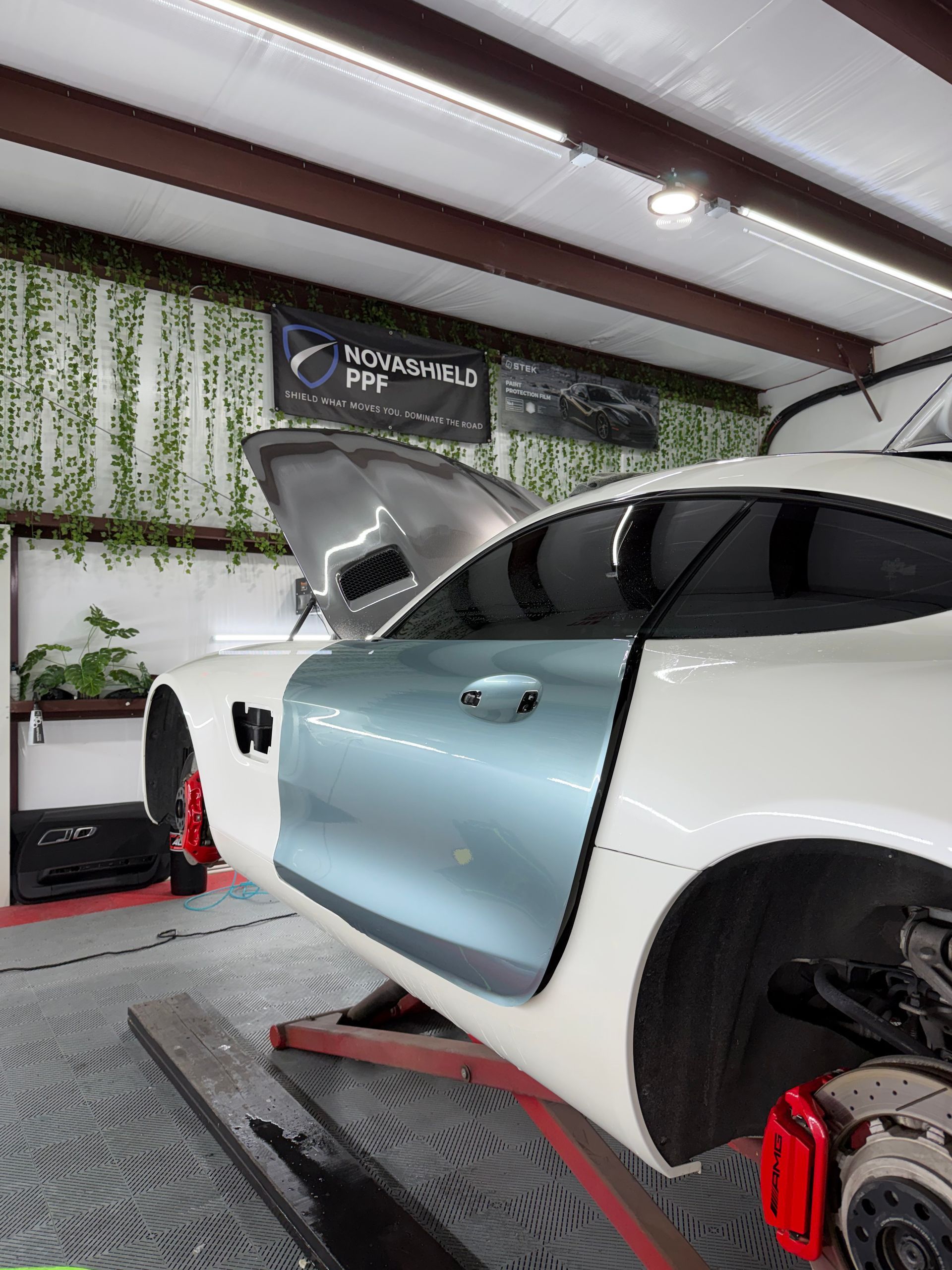 White sports car in a garage, hood open and driver door ajar, with red wheel clamps visible.