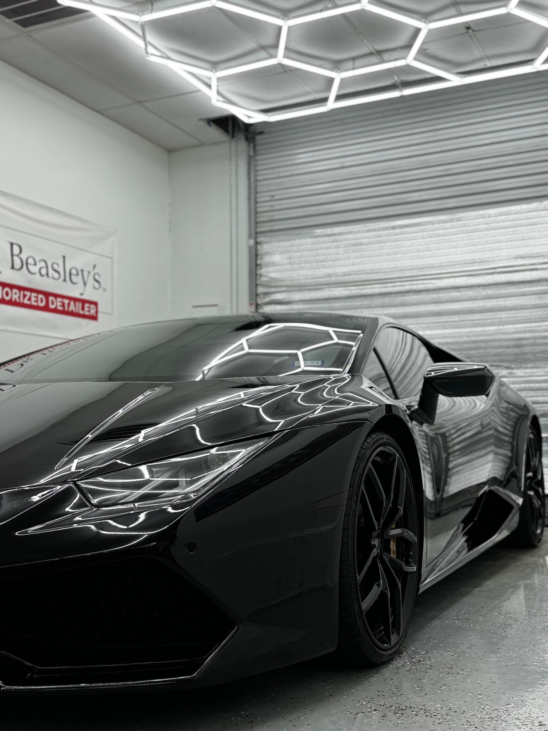 Black sports car in a garage with geometric ceiling lights and a closed roll-up door.