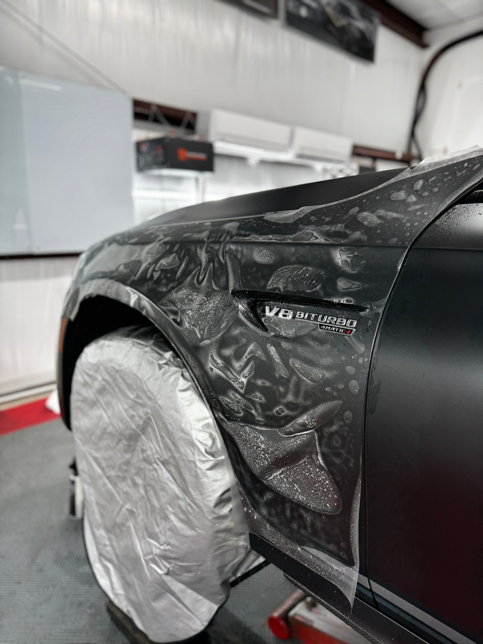 Black car with a silver-covered wheel in a workshop, showing a partially wrapped fender.