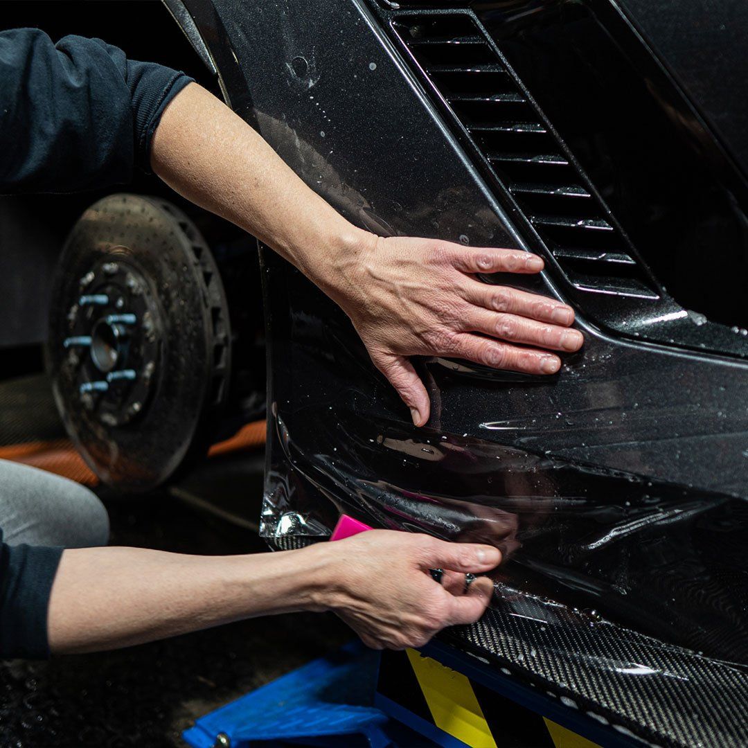 Hands applying black carbon-fiber car body wrap near a wheel in a workshop.