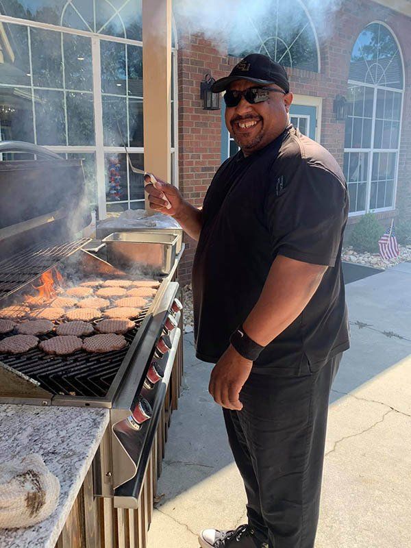 Man grilling burgers outdoors, smiling. Wearing black shirt, hat, sunglasses. Sunny, brick building background.