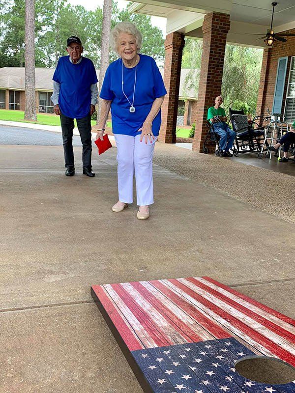 People playing cornhole on a patio; one throws a beanbag, others watch. Board is painted like an American flag.