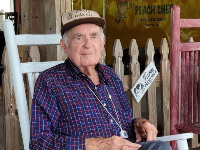 Older man wearing a hat and plaid shirt, seated in a white rocking chair near a Peach Shed sign.