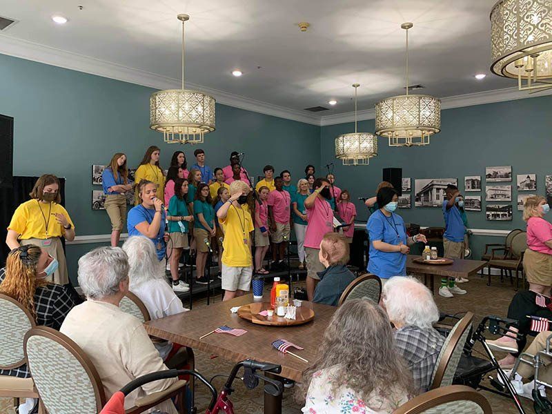 Choir singing to seniors in a room. Performers wear colorful shirts. Seniors sit at tables, some with wheelchairs.