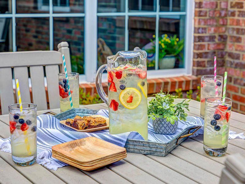 Outdoor table with lemonade pitcher, glasses, and snacks. Red brick background with white-framed window.