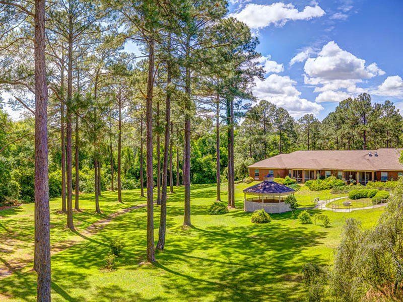 Lush green yard with tall trees in front of a long, brown-roofed house on a sunny day. A gazebo sits nearby.