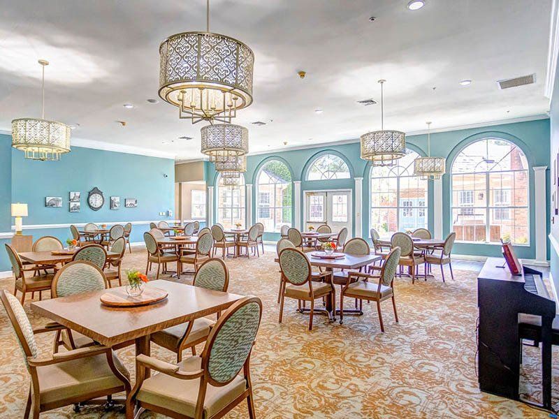 Dining room in assisted living facility; tables, chairs, ornate chandeliers, blue walls, patterned carpet, piano.