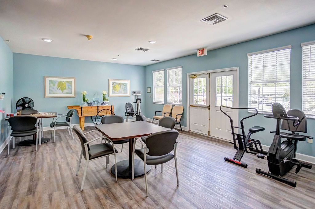 A light blue exercise/activity room with tables, chairs, and exercise bikes by windows.