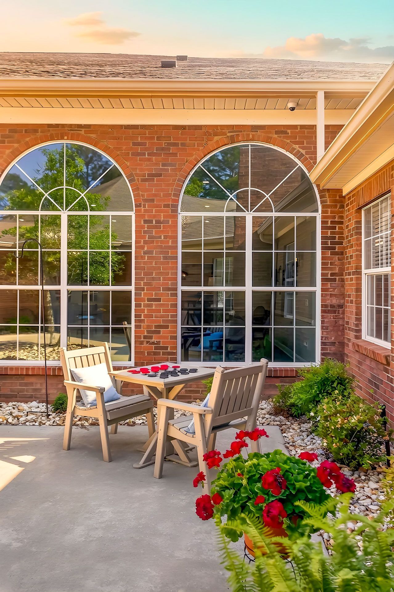 Brick patio with arched windows, wooden table and chairs, and a red flower planter in sunlight