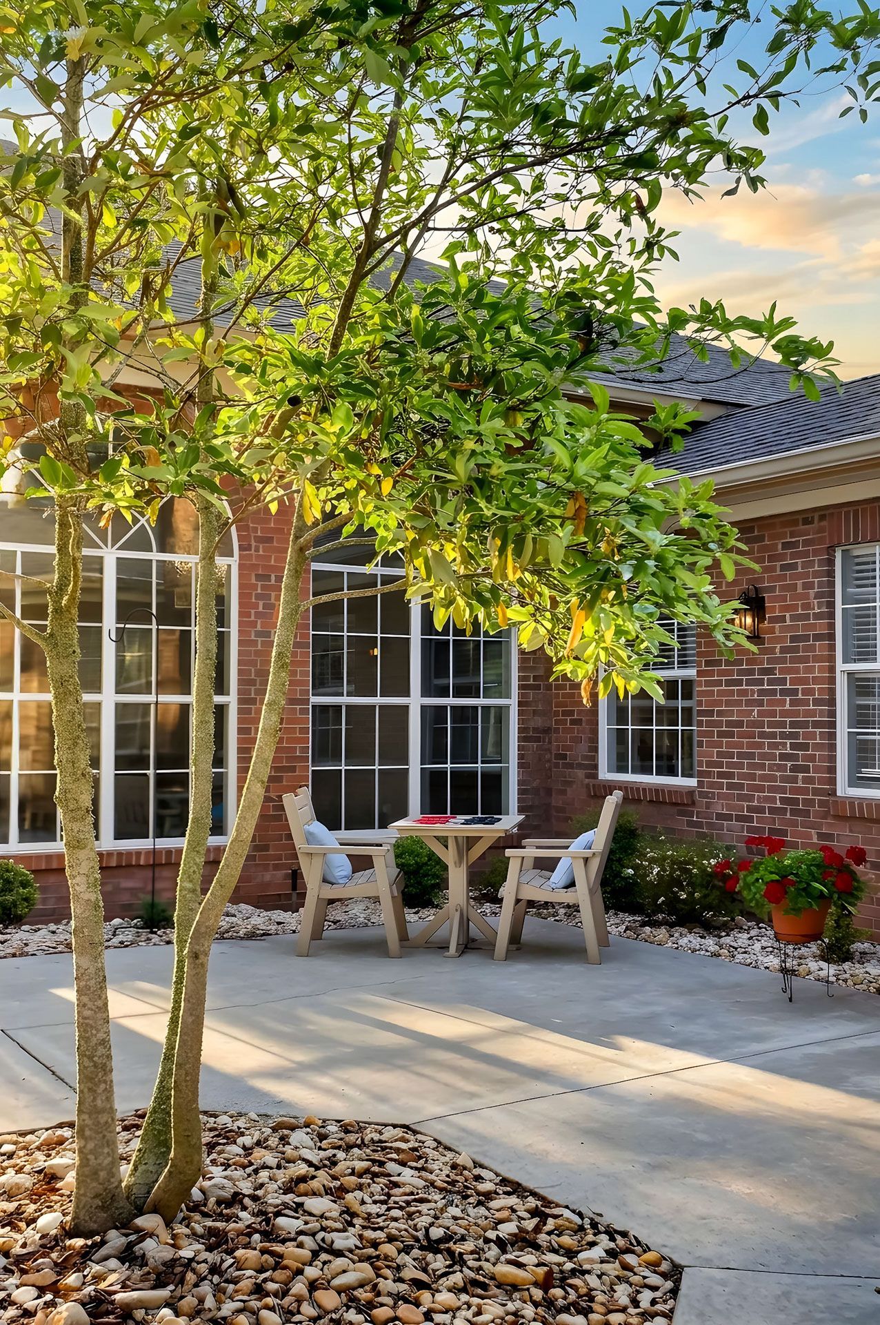 Brick house patio with potted flowers, white chairs, and a leafy tree in front