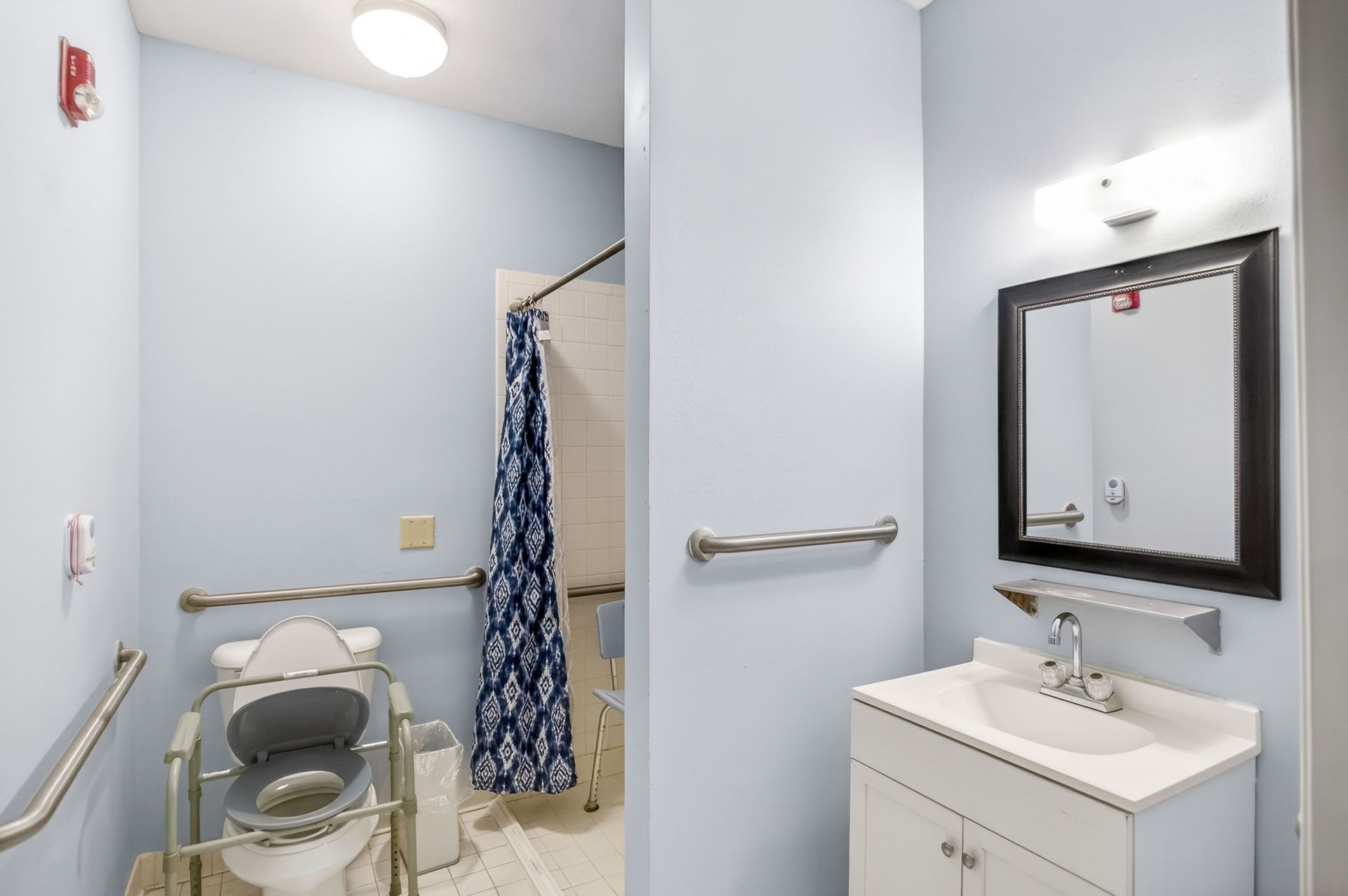 Accessible bathroom with toilet, sink, mirror, grab bars, and shower curtain in light blue walls