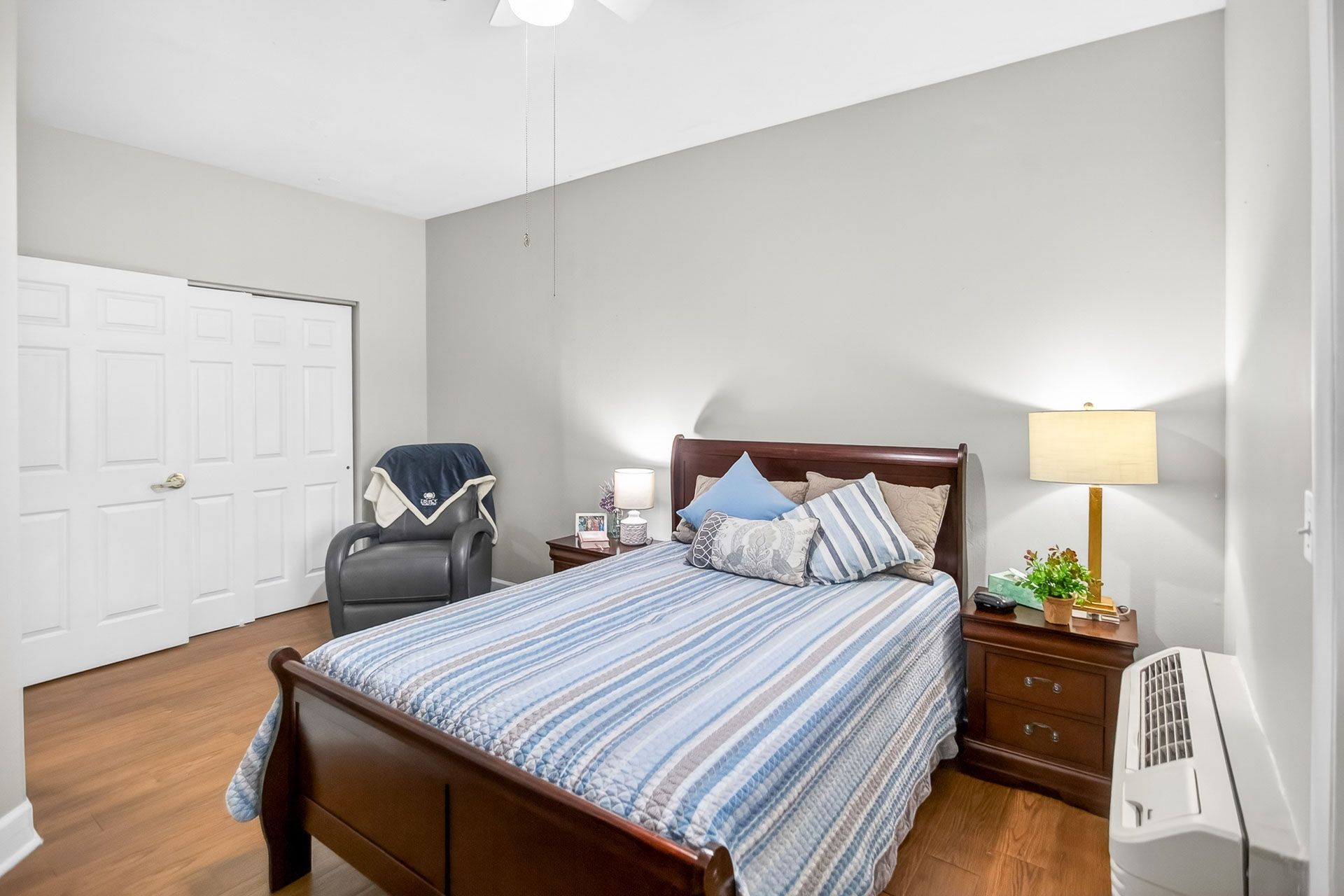 Bedroom with a wooden bed, striped blue bedding, bedside lamp, chair, and white closet doors