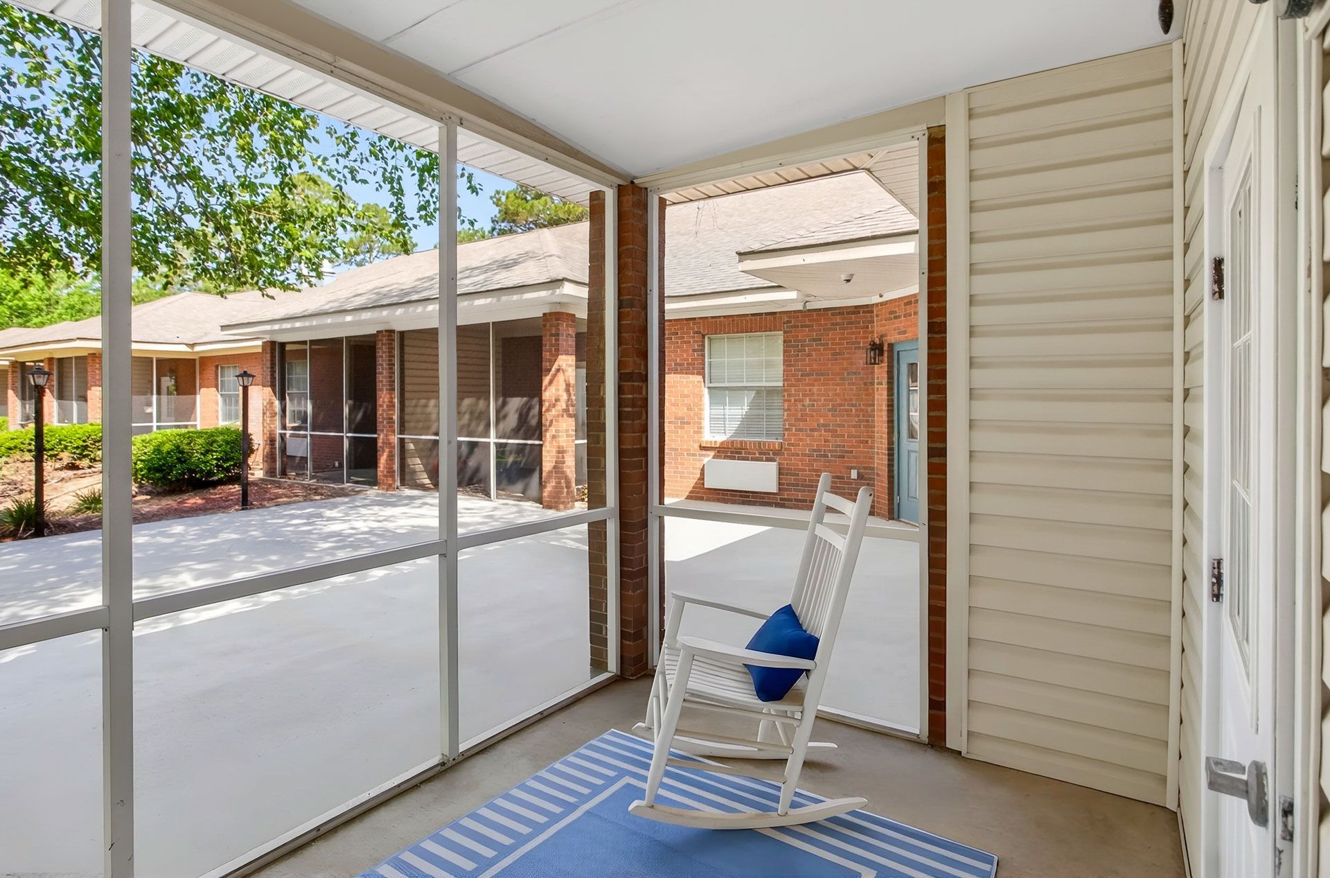 Screened porch with a white rocking chair, blue cushion, and view of a brick apartment courtyard
