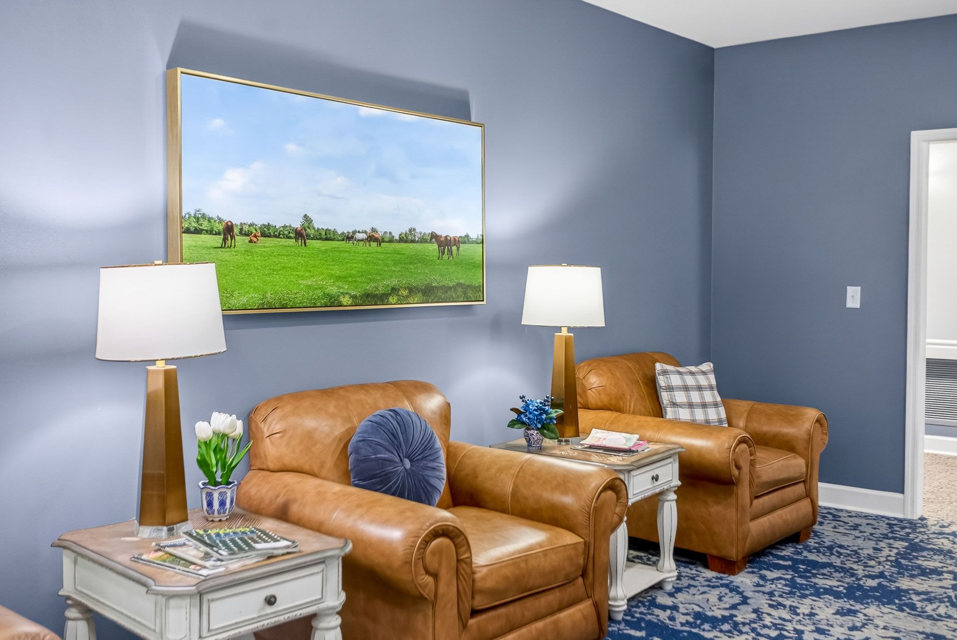 Living room with tan armchairs, blue walls, lamps, and a landscape painting above the seating area