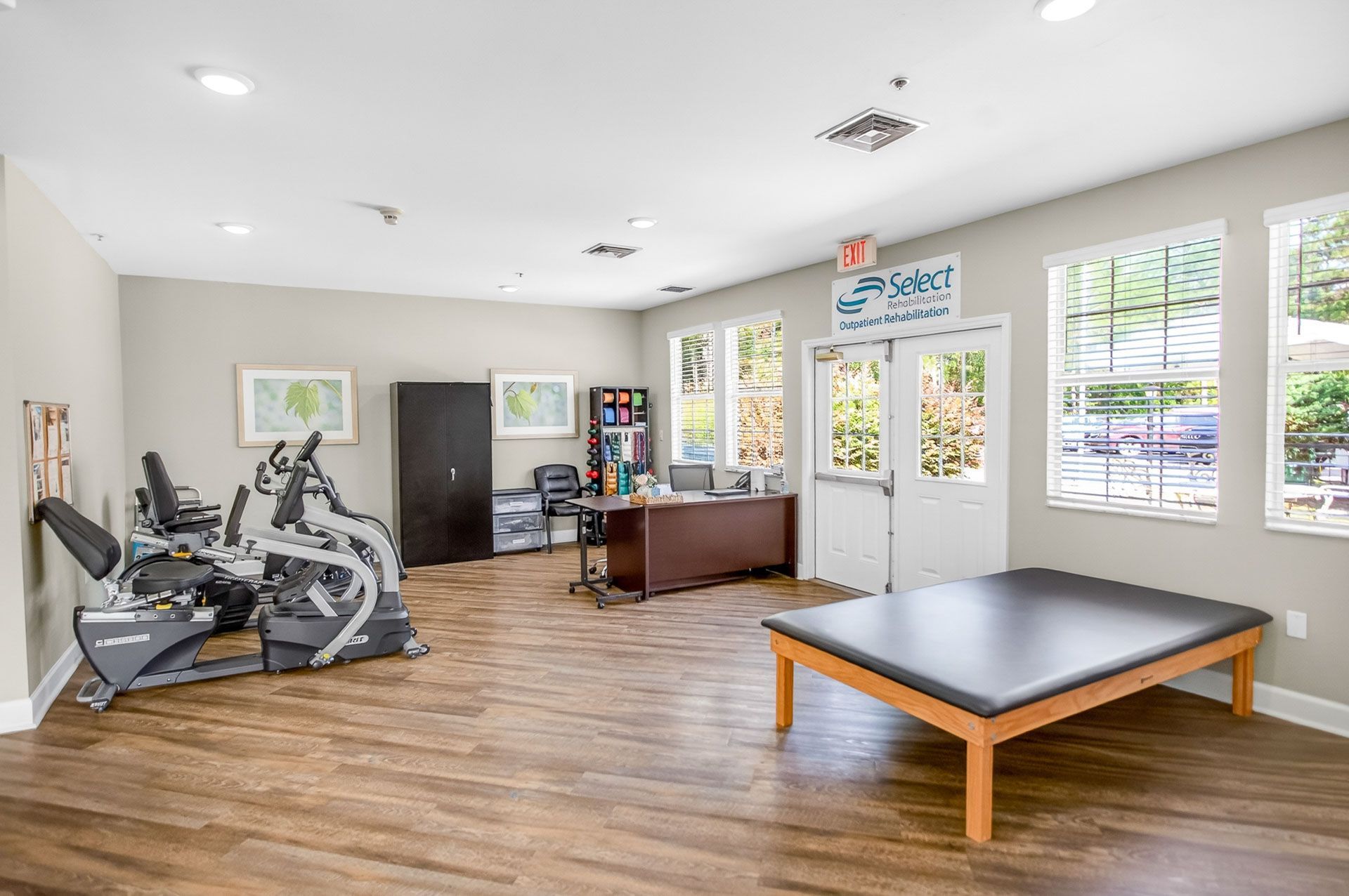 Bright exercise room with gym equipment, desk, windows, and a low table on wood floors