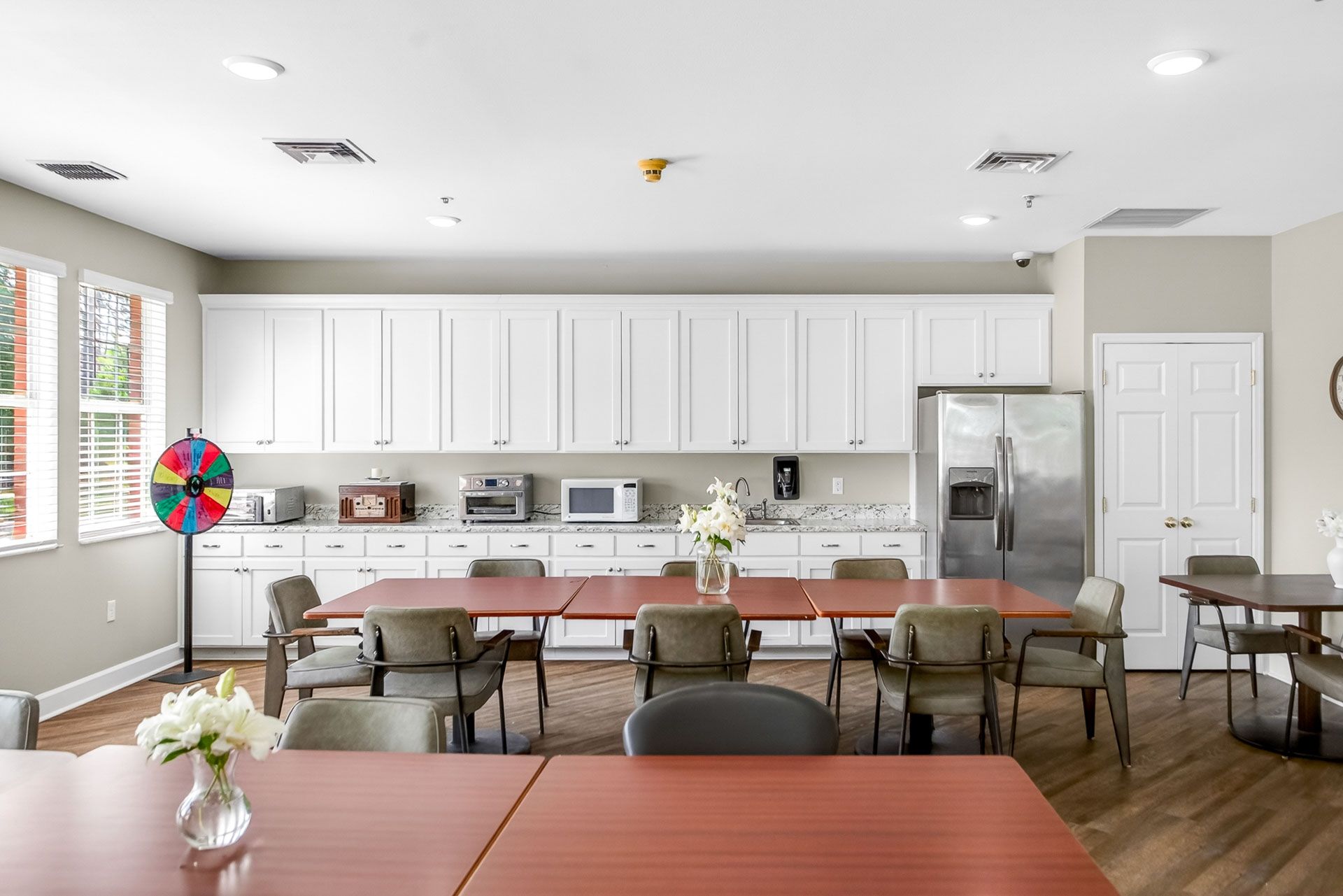 Bright kitchen dining area with white cabinets, tables, chairs, and large windows