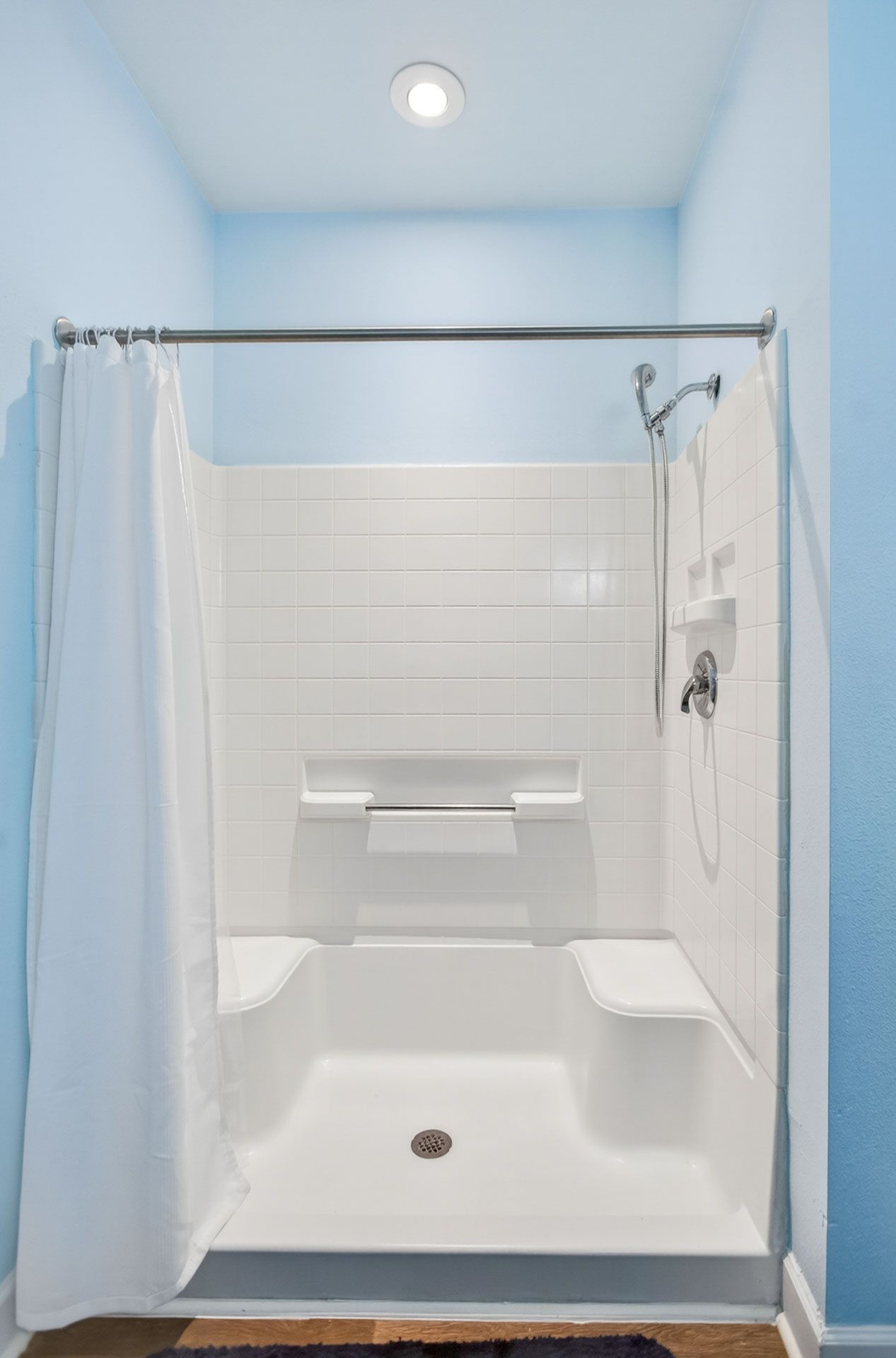White tiled walk-in shower with grab bars, built-in bench, and a showerhead in a light blue bathroom