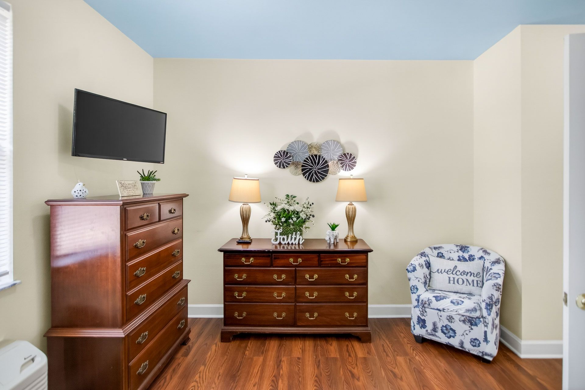 Bedroom with wooden dressers, wall-mounted TV, lamp, flowers, and a blue patterned chair.