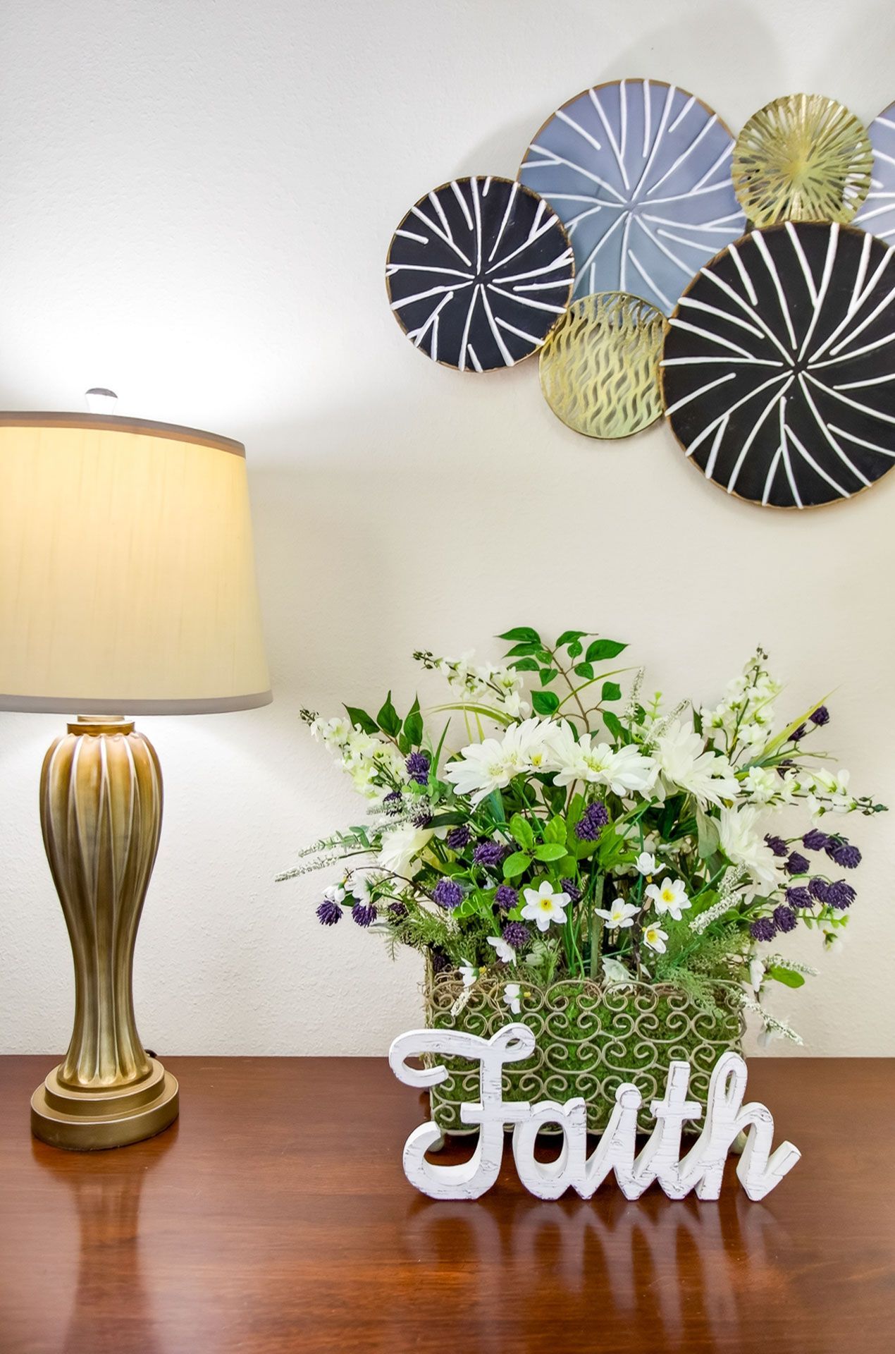 Tabletop floral arrangement with “Faith” sign beside a lit lamp and decorative wall art