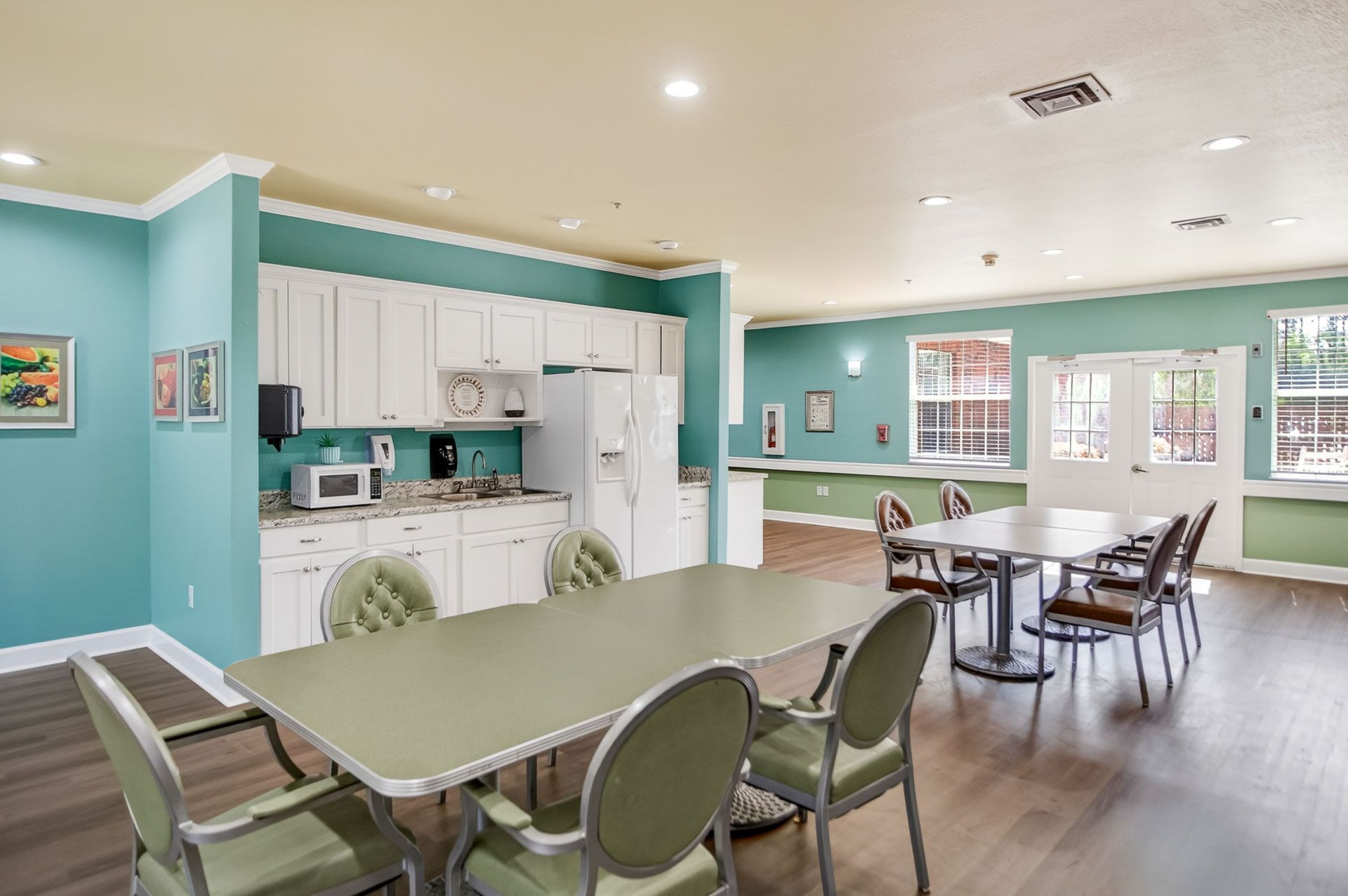 Bright communal dining room with teal walls, white kitchen, tables, chairs, and washing machines.
