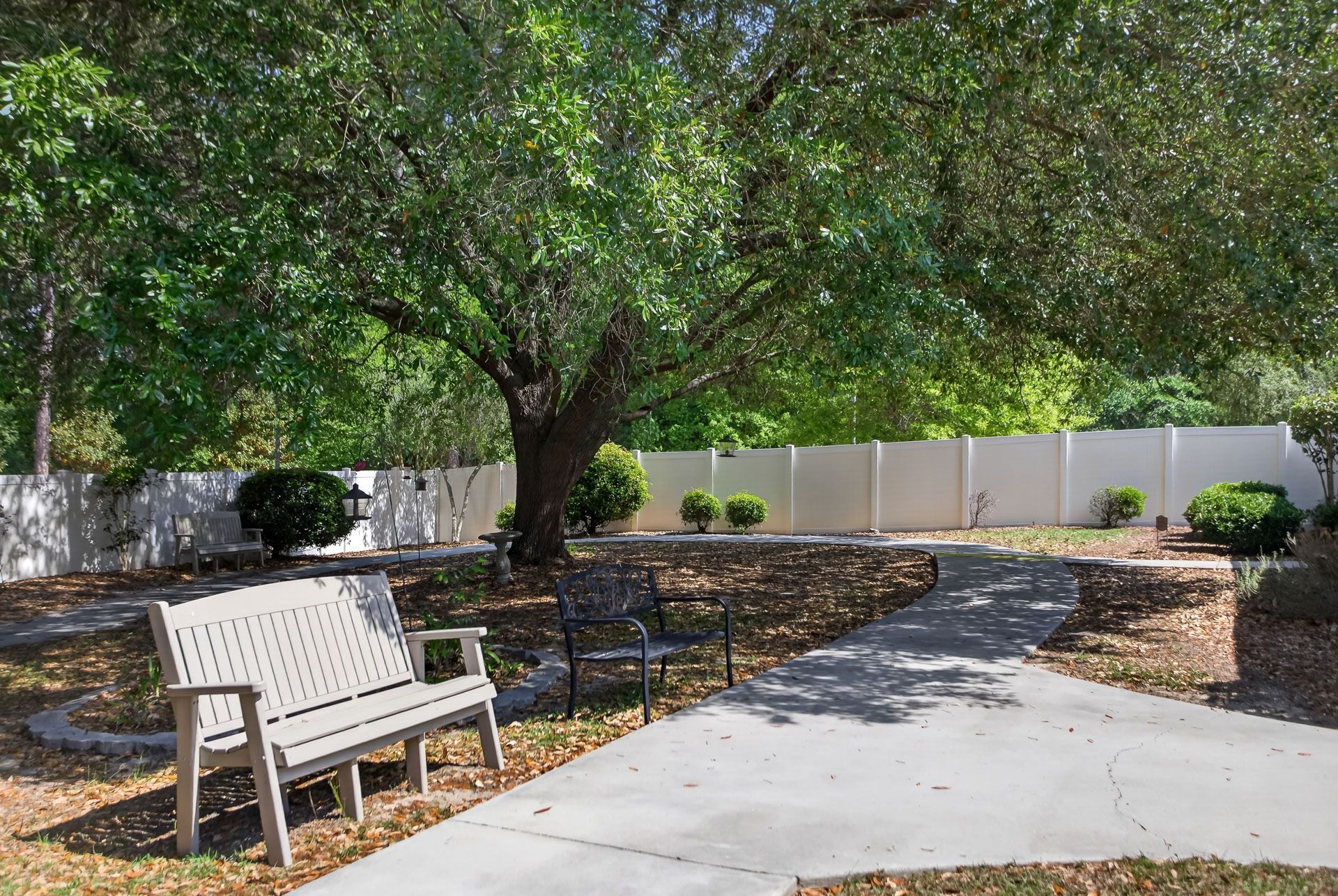 Park bench and winding sidewalk under a large tree in a landscaped courtyard with white fence panels.