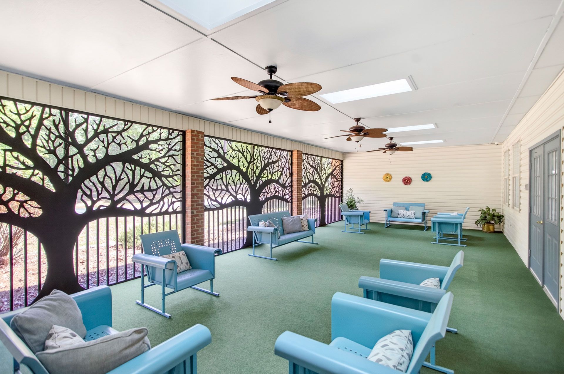 Bright classroom with turquoise chairs, green carpet, tree mural windows, and ceiling fans.