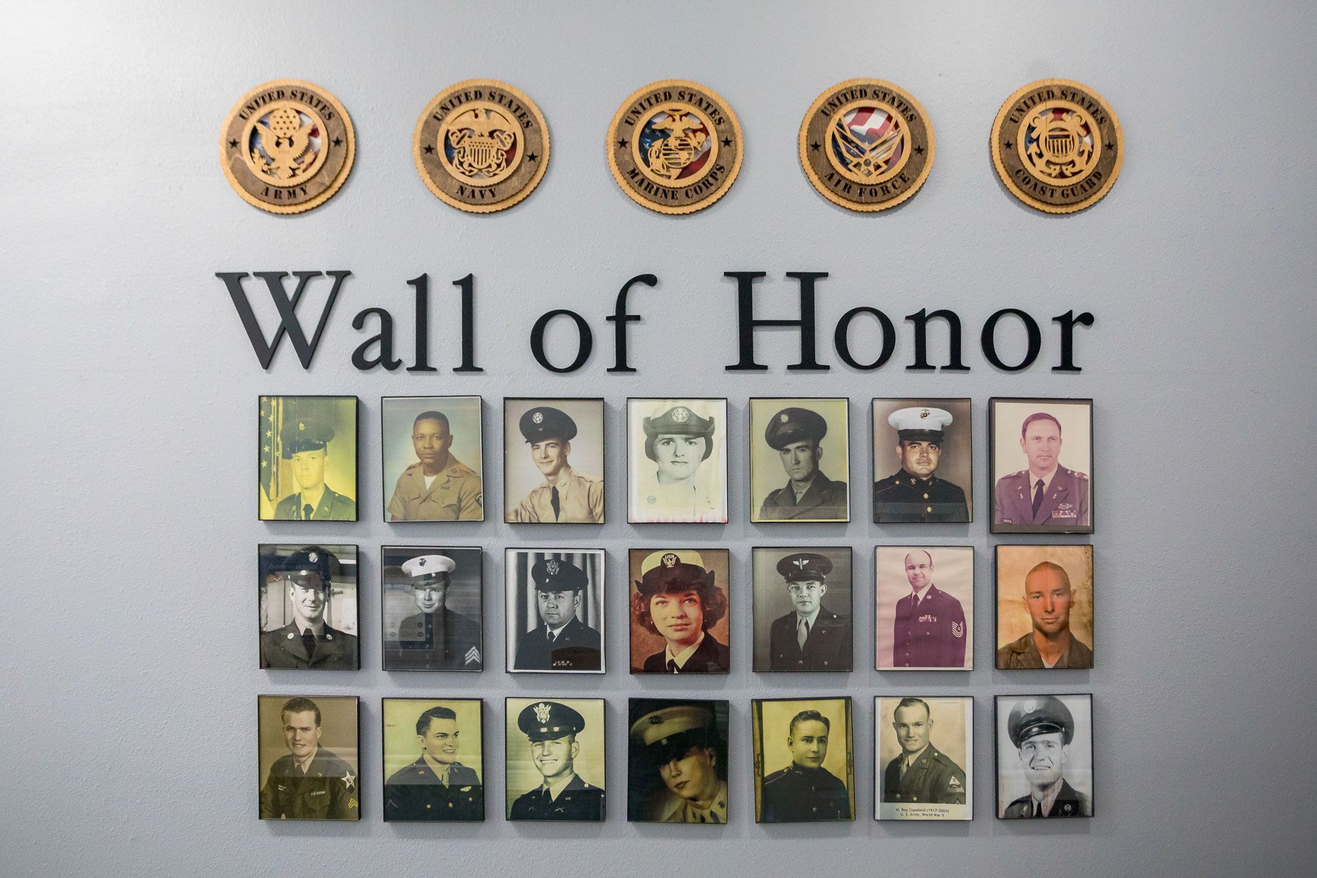 Wall of Honor plaque with five emblems above a grid of framed portraits on a gray wall