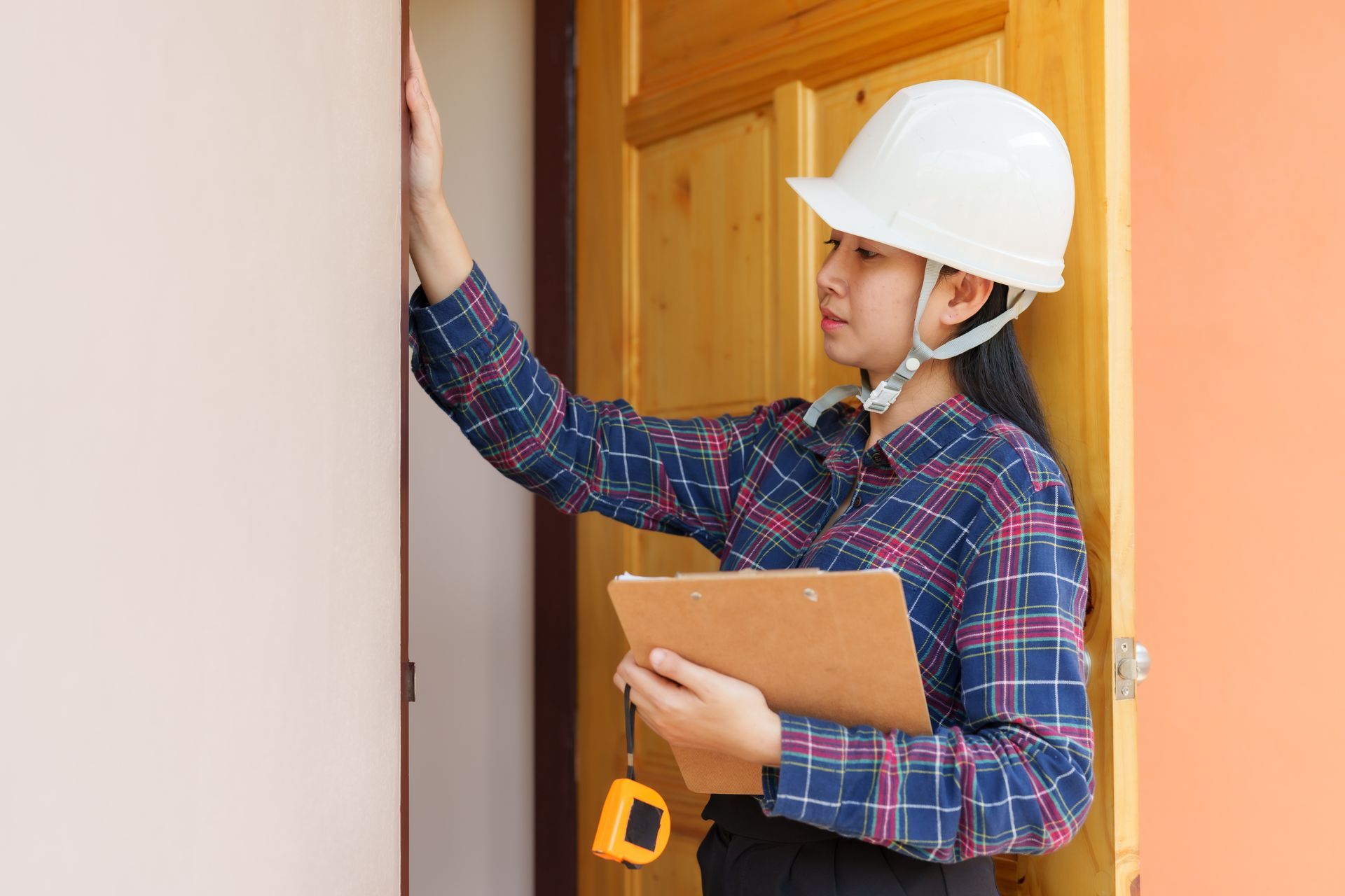 A woman wearing a hard hat and holding a clipboard is measuring a door.