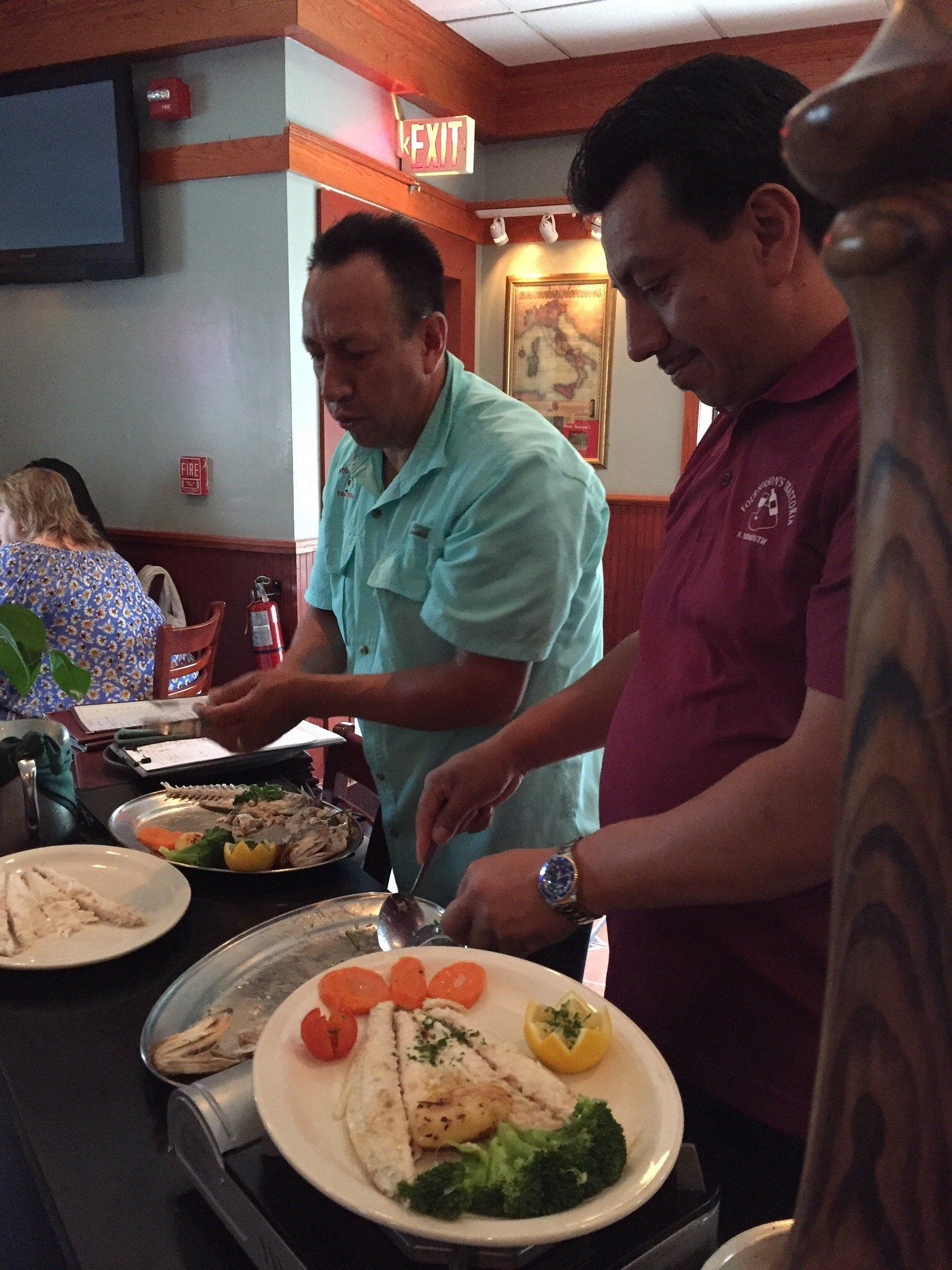 Two men are serving food in a restaurant with an exit sign in the background