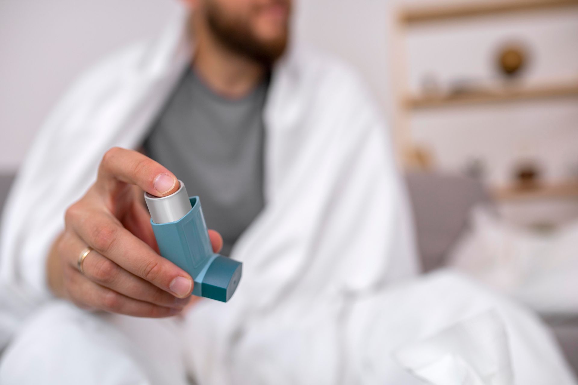 A man wrapped in a blanket holds a blue asthma inhaler as asthma treatment for adults.