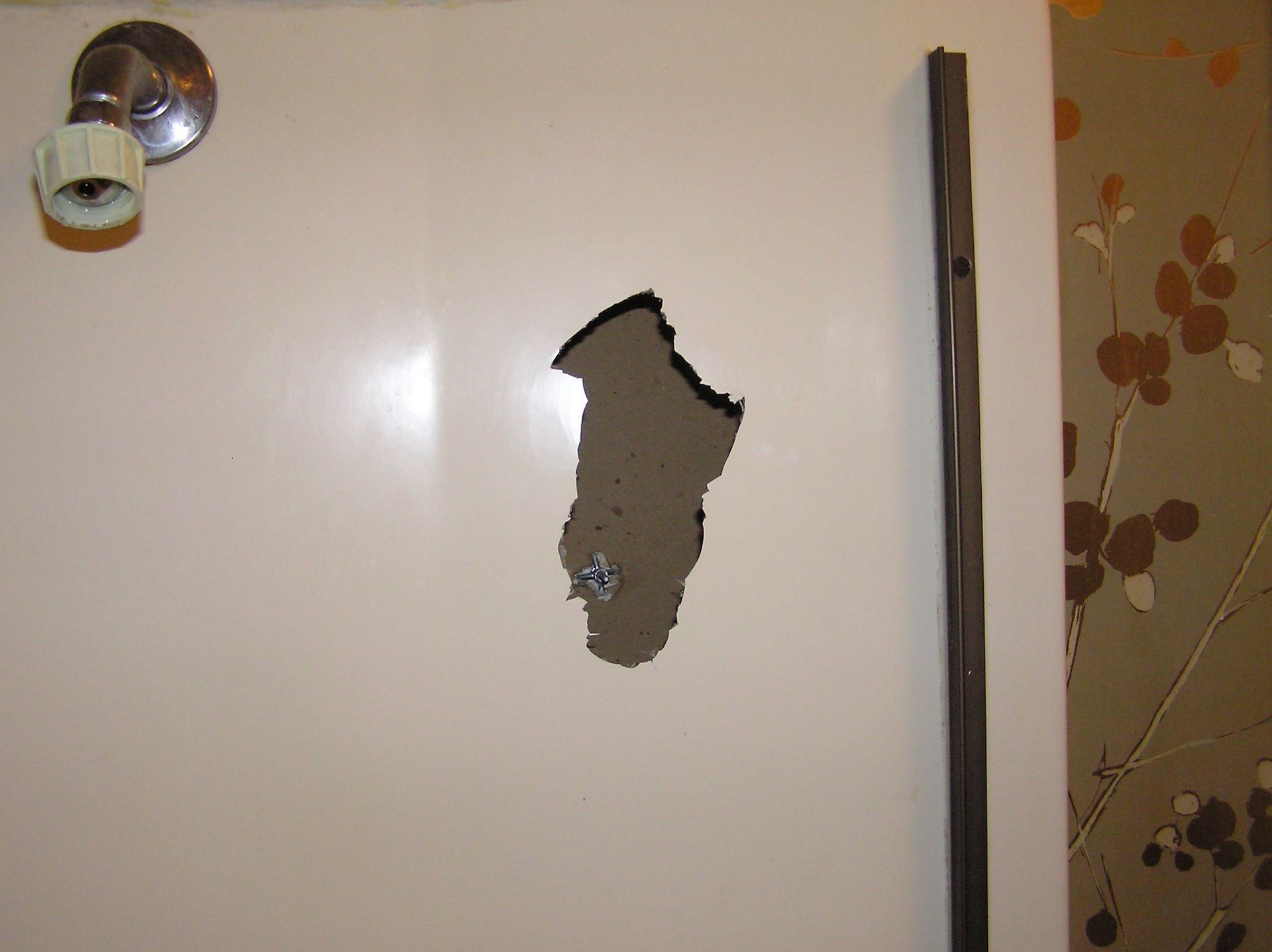 Hole in a white shower wall. Visible water fixture, brown plaster and wall trim. Wallpaper with brown floral pattern.