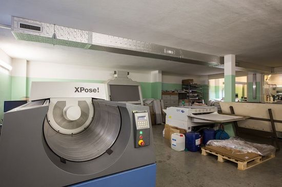 vista di un macchinario da tipografia visto da vicino e sulla destra una macchina da stampa, un bancale di legno con accanto dei prodotti chimici e vicino al soffitto un condotto di ventilazione di metallo
