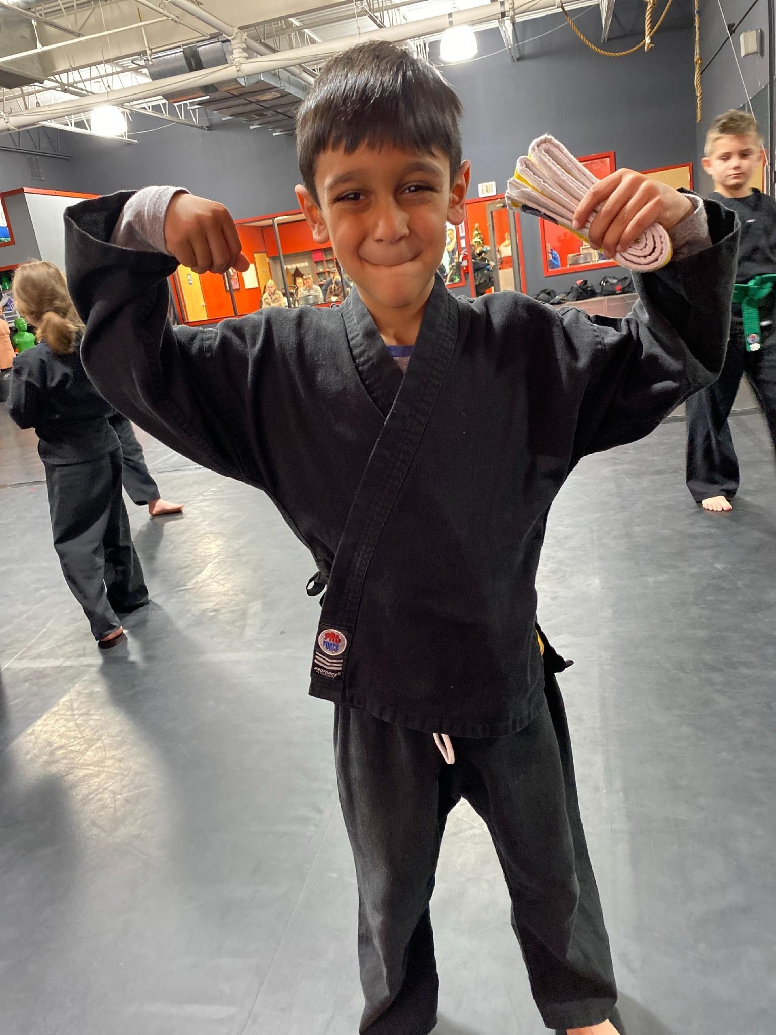 a young boy in a black karate uniform is flexing his muscles in a gym .
