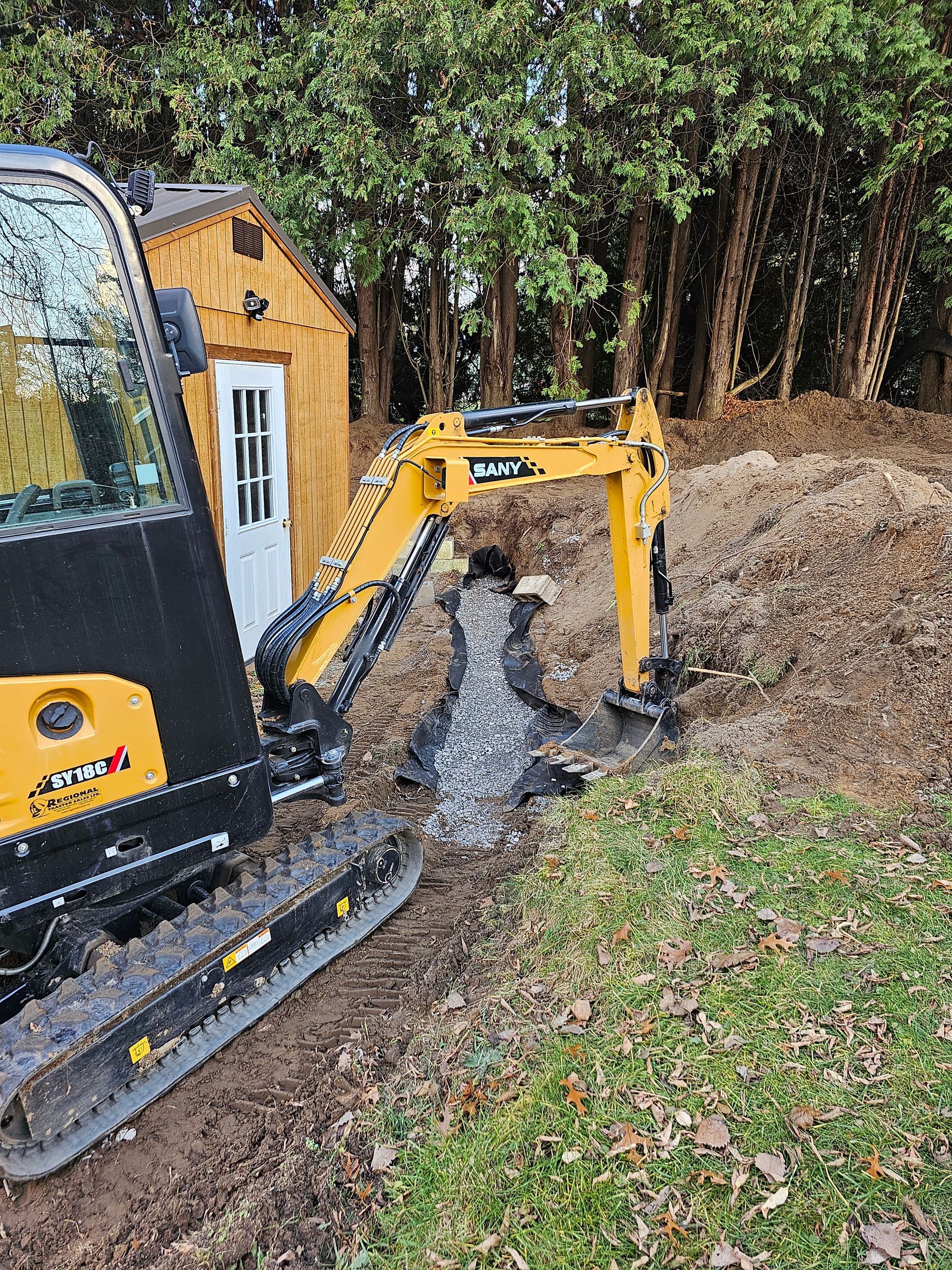 a small excavator is digging a hole in the ground in front of a wooden shed .