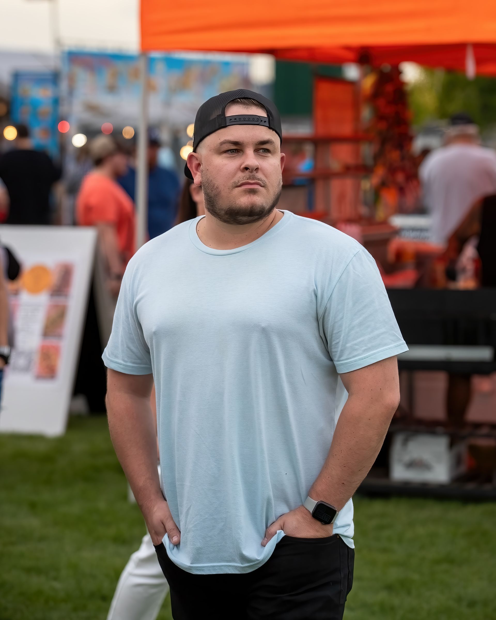 A man wearing a baseball cap and a light blue t-shirt is standing in the grass with his hands in his pockets.