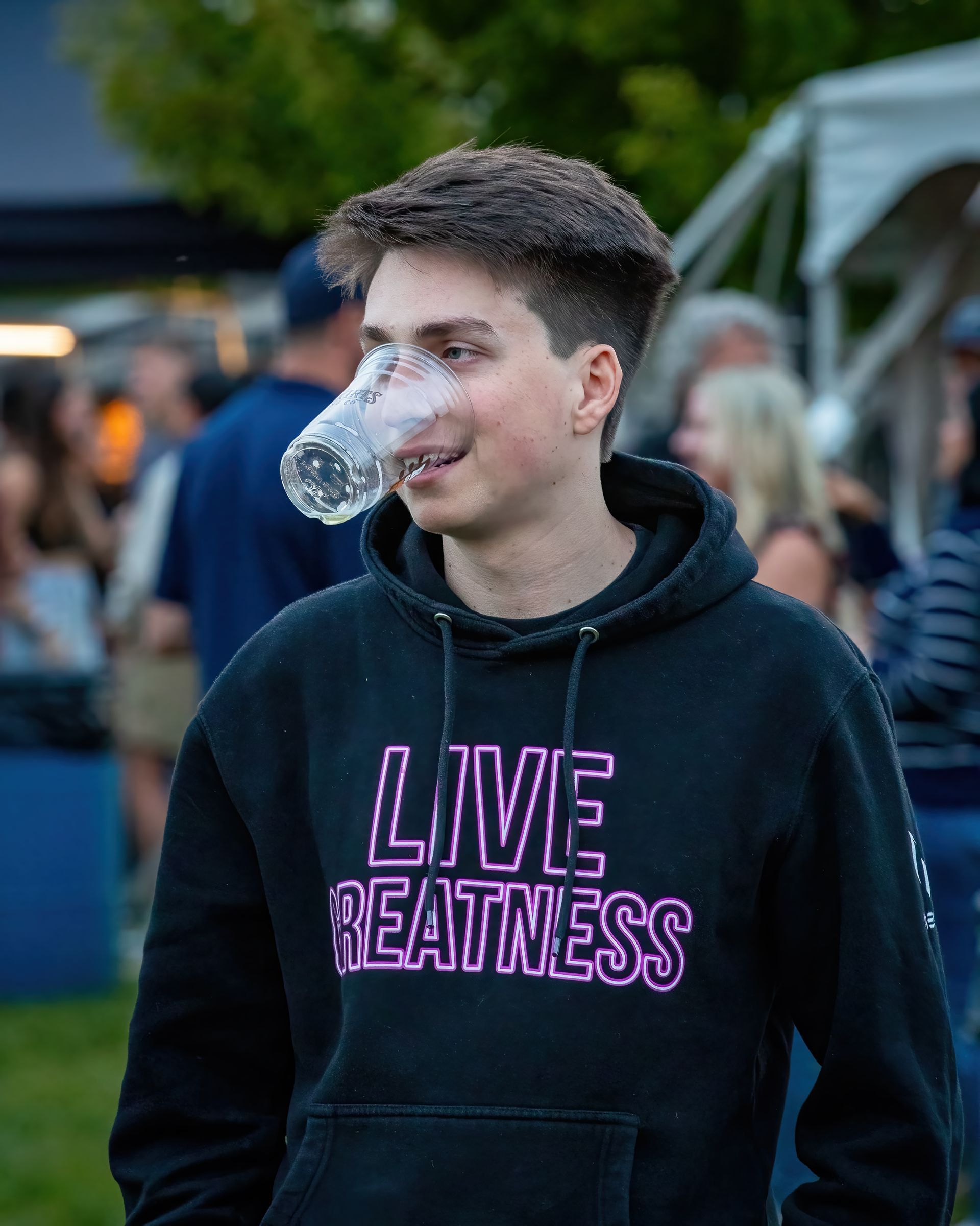 A young man wearing a live reatness hoodie is drinking from a plastic cup.