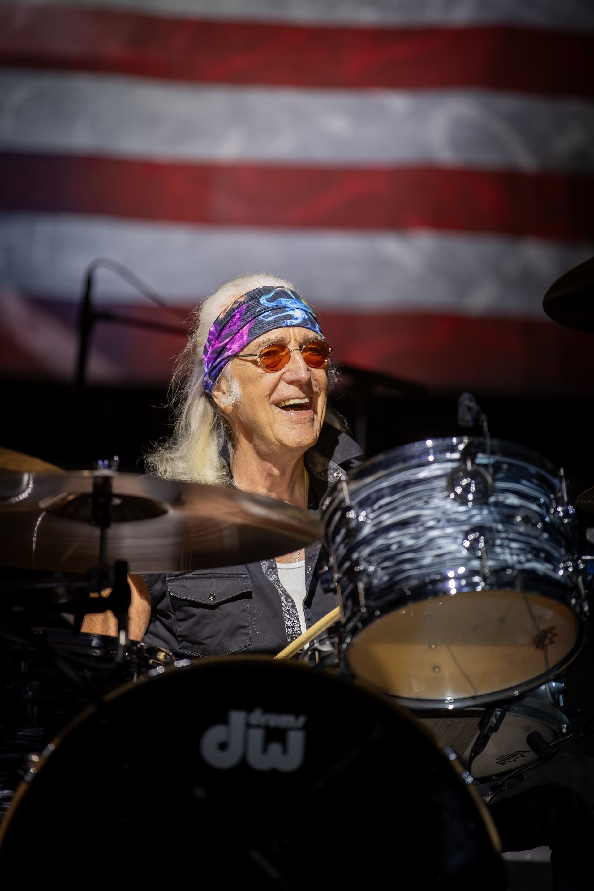 A man is playing drums on a stage in front of an american flag.